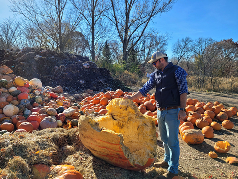 Man standing beside a piece of a large pumpkin with other pumpkins and compost in the background.