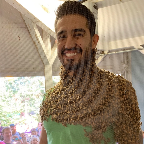 Josh smiles while he is covered from beard to chest with honey bees. He appears to be at a fair with an audience.