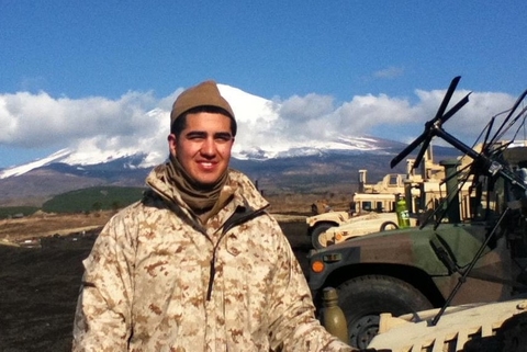 Josh is in brown and tan camouflaged jacket standing next to military vehicles and a snow covered mountain behind him on a clear day 