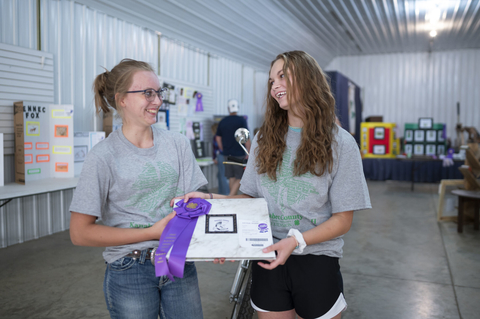 Two teen girls smiling at each other while holding a book that has a purple champion rosette ribbon on it.
