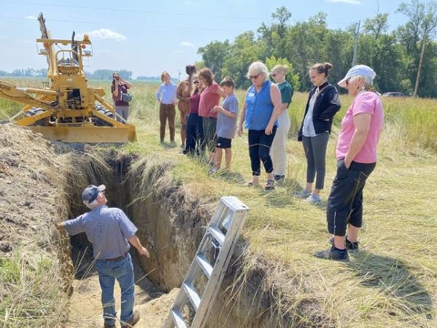 Man standing in a long trench soil pit in a field talking to a group of adults standing on the ground above the pit.