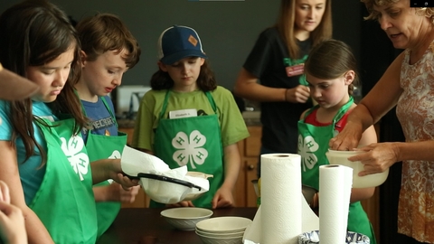 four youth, one adult volunteer and one young adult helper make cheese. The youth are wearing 4-H branded green aprons. A girl in the foreground is straining the cheese through a strainer with a cloth.