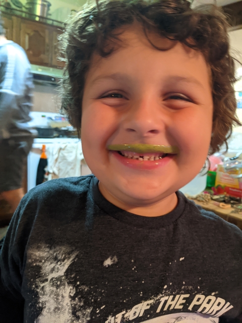 Smiling curly-haired boy shows off his green mustache. He is in a kitchen.
