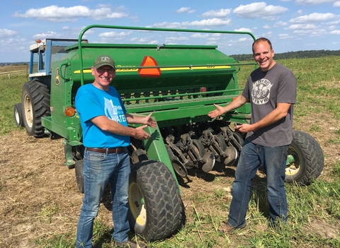 Alan Kraus and Kaleb Anderson standing behind a John Deere seed planter behind a tractor.