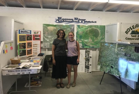 Virajita Singh and Miranda Olson standing together in a booth full of images of the Roseau River recreation project.