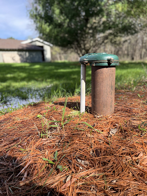 Private well pipe and cap emerging from the ground with standing water in the background.