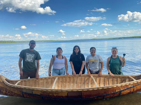 A group of youth standing behind a Birchbark Canoe next to a body of water.