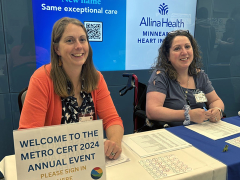 Anna Peterson (right) sitting in a wheelchair behind a table; alongside fellow CERTs regional coordinator, Heidi Auel. A sign at the table welcomes visitors to the “Metro CERT 2024 Annual Event.” Both women are smiling, welcoming guests to the event.