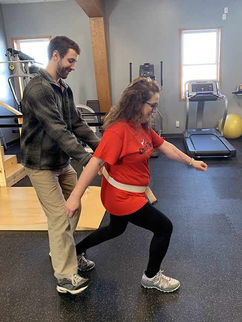 Peterson taking part in physical therapy. She stands in a lunge position while Trevor Roppel, DPT of Choice Therapy stands behind her, helping support her. Exercise equipment stands in the background.