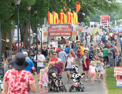 A crowd of people enjoying a county fair, with a corndog and hotdog stand in the background.