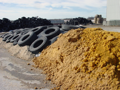 Plastic-covered pile of distillers grains weighted down by tires, next to an uncovered pile.