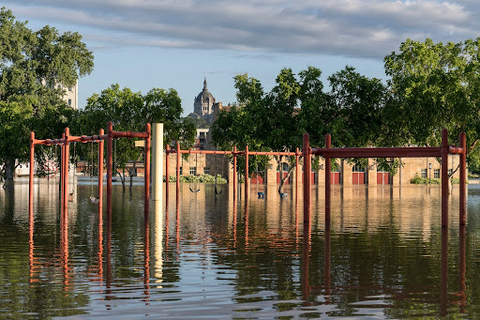 A children's playground with swings and climbing structures in a flooded area with trees and cityscape in the background.