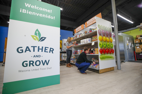 A large welcome banner for Gather and Grow food shelf next to two people stocking shelves