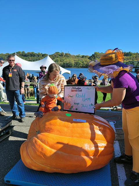 Woman with baby posing by a giant orange pumpkin with another woman holding a sign that reads: Stillwater Harvest Fest. Grower: Klodd, U of M. Weight: 744.5 pounds.