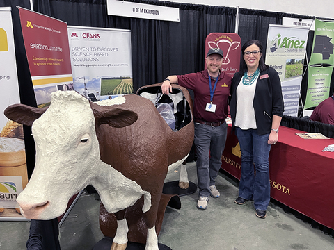 Two people stand next to a life-sized model of a cow at a tradeshow booth. The cow's back panel is removed to show the interior of the cow.
