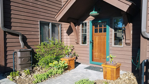 The front entry of a brown, wood house with shrubs and flowers under a window and a brown rectangular rain barrel positioned under a downspout.