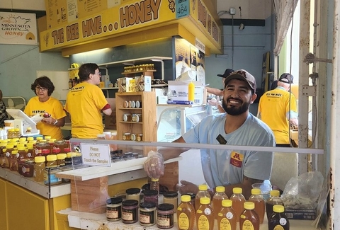 Josh smiles in front of many jars of honey. There is a honey sign behind him and a Minnesota Grown sign on the wall. 