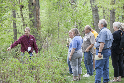 People standing in a wooded area listening to someone speaking and pointing out plants.