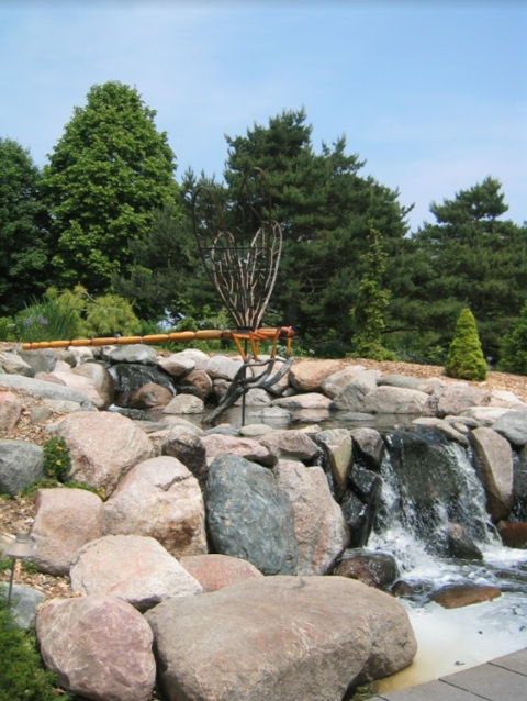 A water feature consisting of large boulders and an artwork with large dark green evergreens in the background,