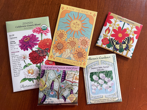 Various seed packets decorated with colorful art lying on a wooden table.