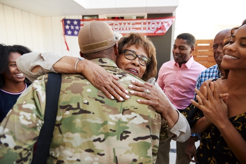 An elderly woman hugs a man in soldier fatigues with a welcome home banner and smiling loved ones looking on. 