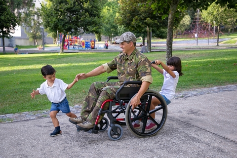 A smiling man in soldier fatigues and using a wheelchair plays with two young children in a park. 