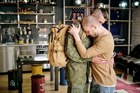 Two men embrace in a kitchen, one is wearing a soldier's uniform. 