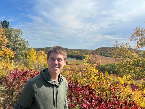 Teen in olive green hooded sweatshirts stands outdoors with a blue sky, yellow and red fall foliage, and hills behind him.