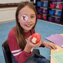 Fifth grade girl holds up an apple with a bite out of it in a classroom. She has a sticker over one eye that shows an apple and says "Crunch"