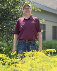 Brian stands in a field of small yellow flowers in front of the plant growth building on campus.