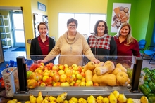 Four educators stand behind a bin of apples and squash in a colorful, well-lit food shelf 