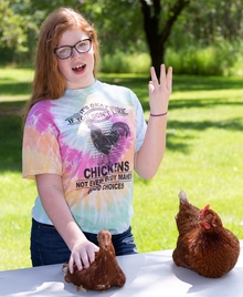Teen presents to a video camera, which is out of frame, about her two chickens that are on the table in front of her.