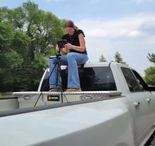 Allison sits on a platform in a pickup truck bed with her phone camera and a tripod, the horse trailer out of frame.