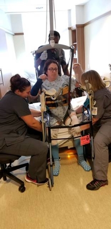 Anna sitting on hospital bed with three therapists around her as they prepare to stand her up to try using a walker.