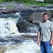 Brent Mason stands on rocks beside a rapid river with some waterfalls.