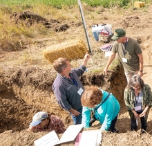 Six people in a trench as field day. Two are talking to each other and the others are chiseling soil.