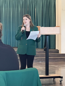 Victoria presenting in front of lectern, holding a piece of paper and wearing her green 4-H ambassador blazer speaking into a microphone.