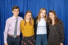 Four youth stand arm-in-arm, smiling, against a curtain backdrop. 