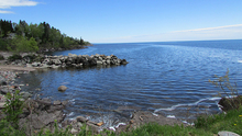 Large body of water with grassy shore in front and rocky shore on left with big hill past the rocks.