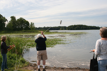 Three people on lake shore; one tossing rake into lake to retrieve samples.