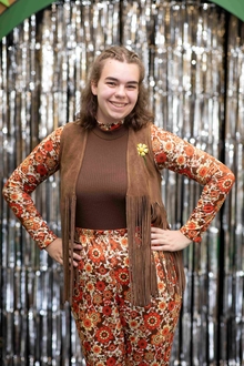 Participant in hippy 70s outfit with fringy vest poses with hands on her hips in front of a glittery background