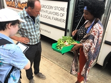 A woman steps out of a bus to show people a tray of fresh veggies