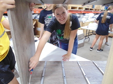Victoria Shafer is working on some sort of woodworking project and smiling at the camera from behind a box that she is working on using a screwdriver