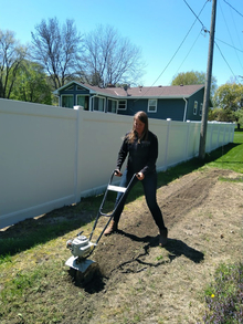 Woman using a rototiller to till long strip of ground in a yard.