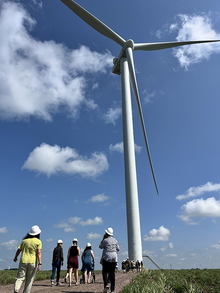 A wind turbine against a blue sky. People walk below it, looking up.