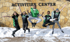 Five teens wearing green 4-H capes jumping in the air outside.