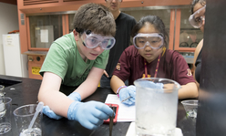 two teens wearing googles and looking at a beaker