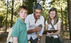 A man showing a pinecone to two kids in a wooded area.