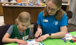 A teen girl wearing a 4-H shirt helping a younger girl with a craft.