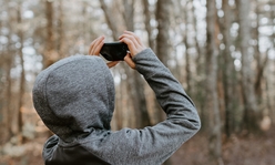 A boy wearing a hooded sweatshirt holding up a phone to take a photo out in nature.
